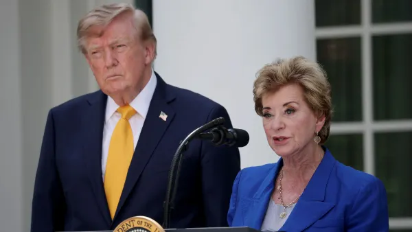 A person with a short haircut in a suit and gold tie stands to the left of a person also with a short haircut wearing a blue suit who is speaking into a microphone