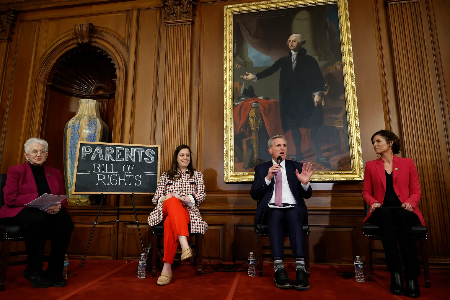 Four adults are seated on a stage in a wood-paneled room. Next to them is a stand with a chalkboard sign that reads "Parents Bill Of Rights."