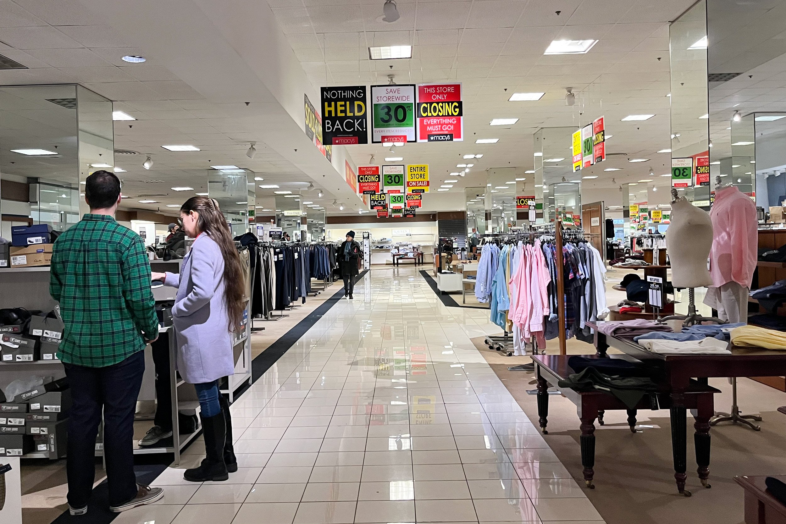 Shoppers walk through a Macy&amp;#x27;s department store as sale signs hang from the ceiling.