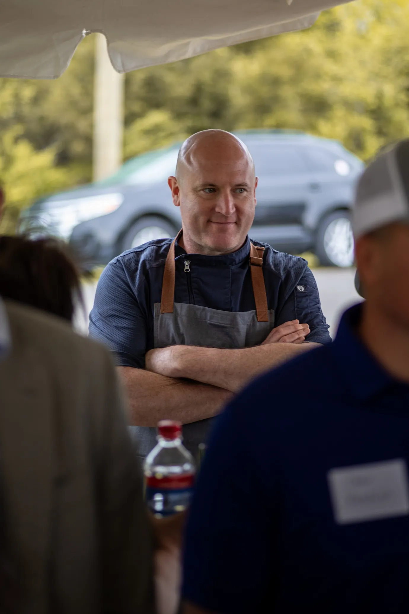 A person stands outside under a tent, wearing an apron, while other people stand indistinct in the foreground.