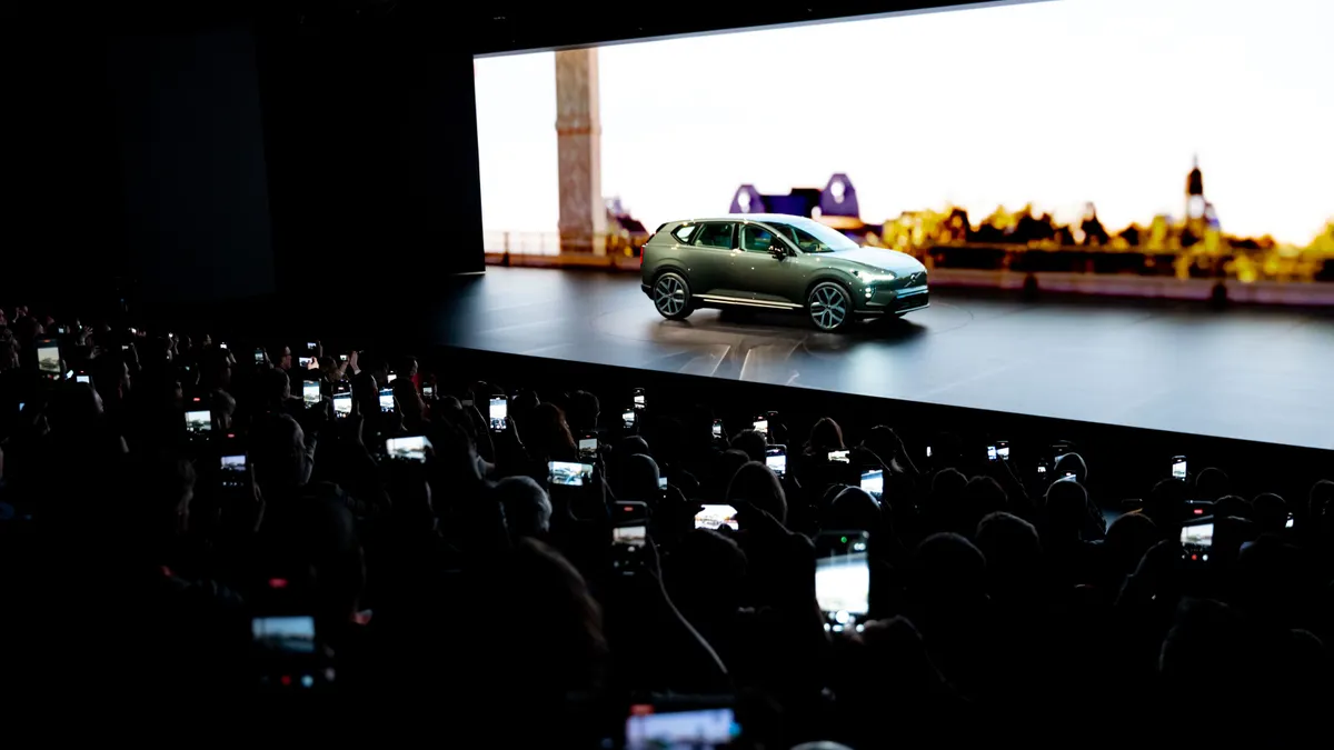 A Volvo EX60 is shown on stage during a reveal event, with the lettering EX 60 on a screen behind it.