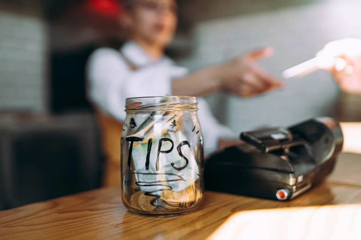 An image of a tip jar in the foreground with a customer paying by credit card in the background