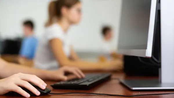 A blurred photo of a teenage student in glasses learning computer science while using a computer in a classroom.