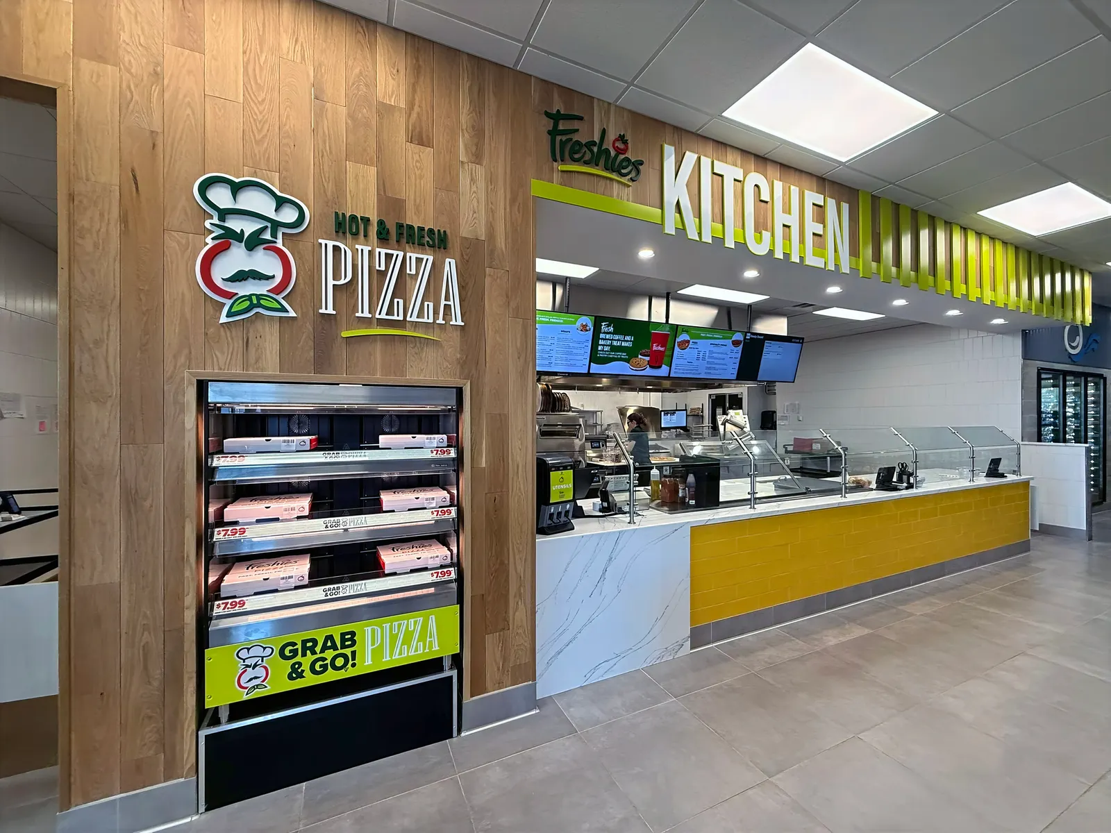 A kitchen area inside a store. A sign over the counter says Freshies Kitchen while a kiosk to the left of the counter holds a number of pizzas in boxes. Text above the kiosk says Hot &amp; Fresh Pizza and a sign beneath it says Grab &amp; Go Pizza.