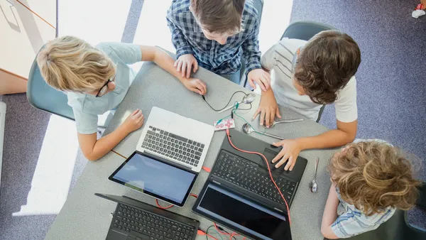 A camera looks down on four students seated at desks, groups together. On the desks are laptops, spoons and wires