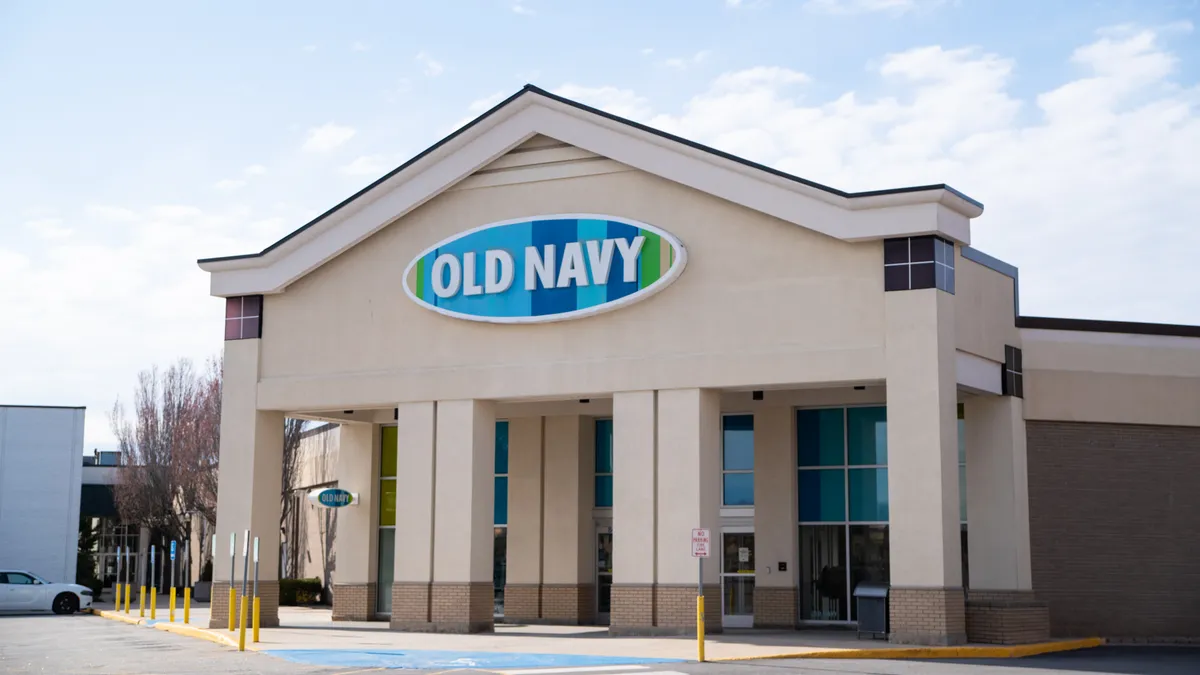 An Old Navy store, with a blue-and-white oval sign on a peaked false front, under a pale blue sky and puffy white clouds.