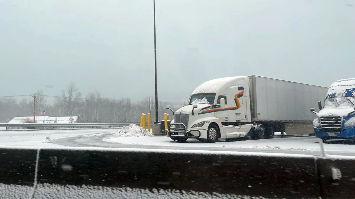 Snow covers windshields of trucks and pavement in a parking area.