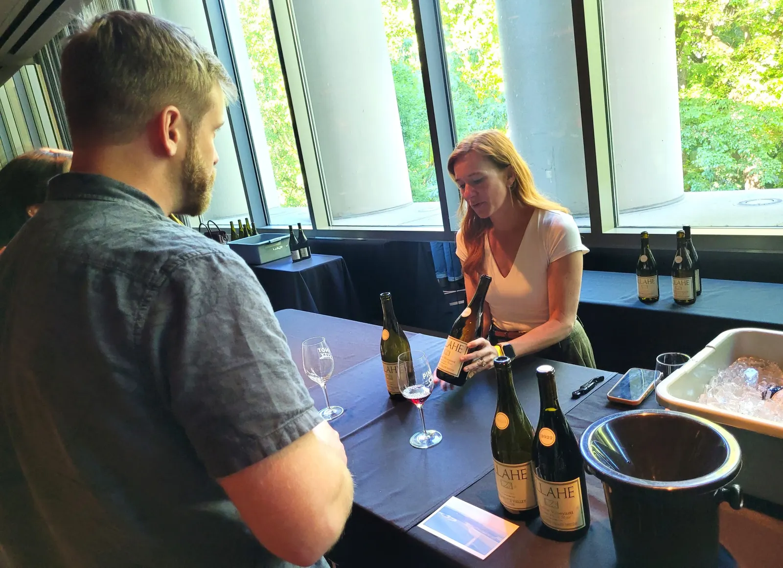 A person behind a table at an event shows bottles of wine to two people on the other side of the table.