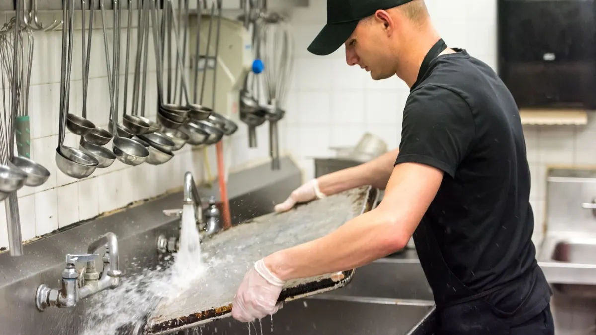 A person wearing black hat and black clothes stands in front of a big sink and is washing a large pan.