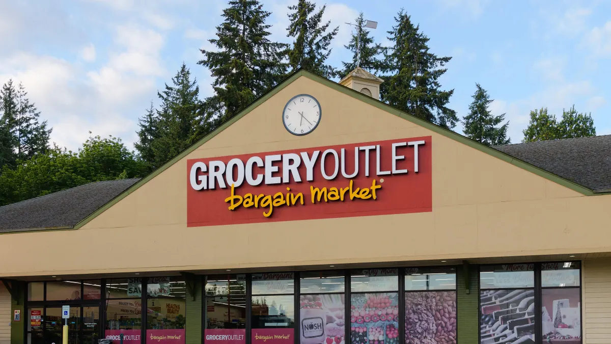 A tan gable-roofed building with a clock, red and yellow “GROCERY OUTLET bargain market” sign, and large window graphics showing produce.