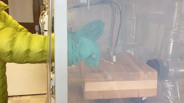 A person wearing a thick yellow coat sticks their arms into large blue gloves and swabs a piece of wood in a plastic container with a Q-tip.
