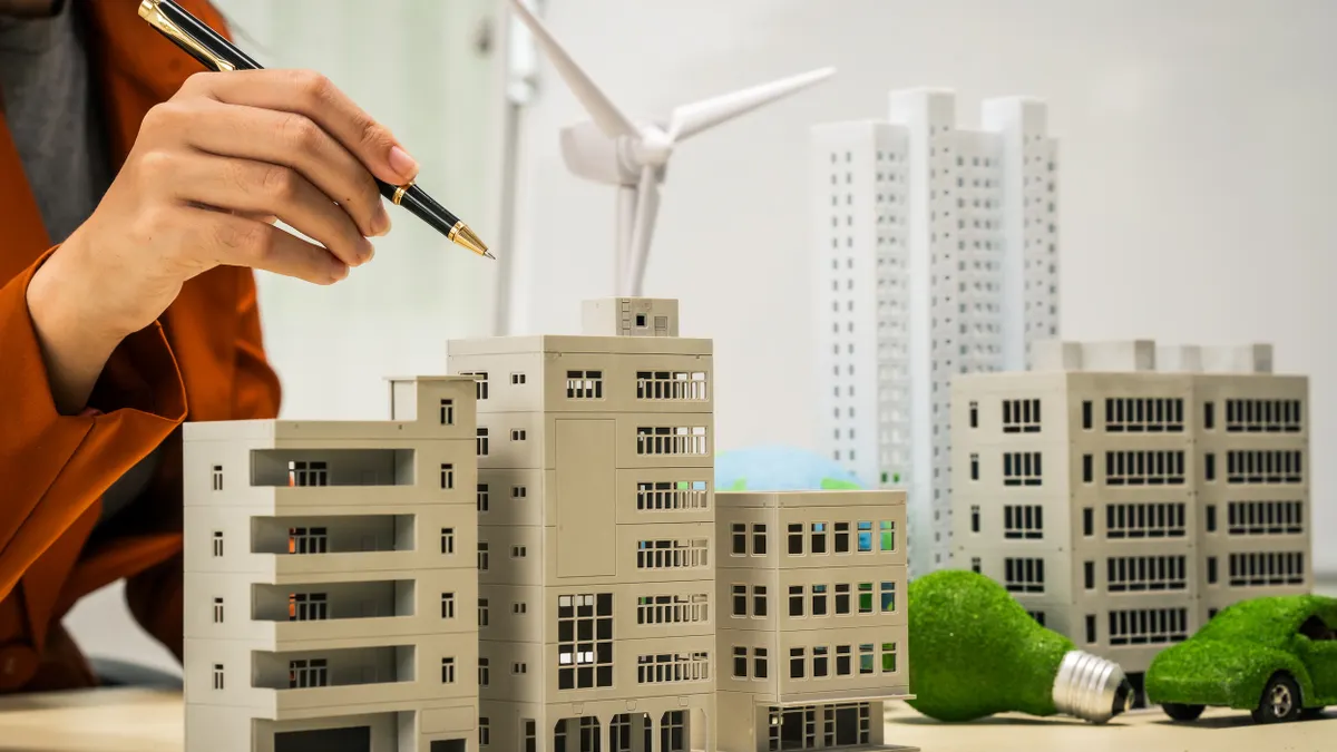 A hand holding a pencil hovers over model buildings and windmills.
