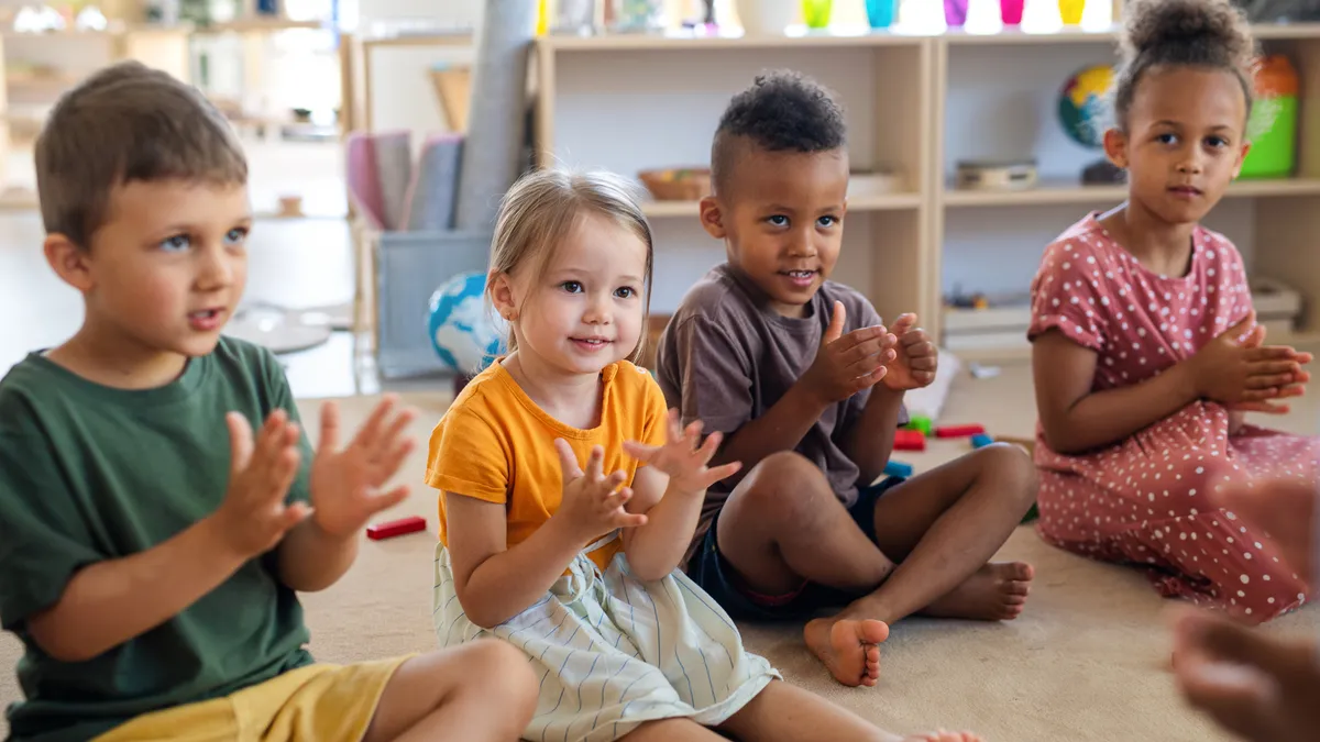 Four young children are sitting on the floor in a classroom. They are clapping