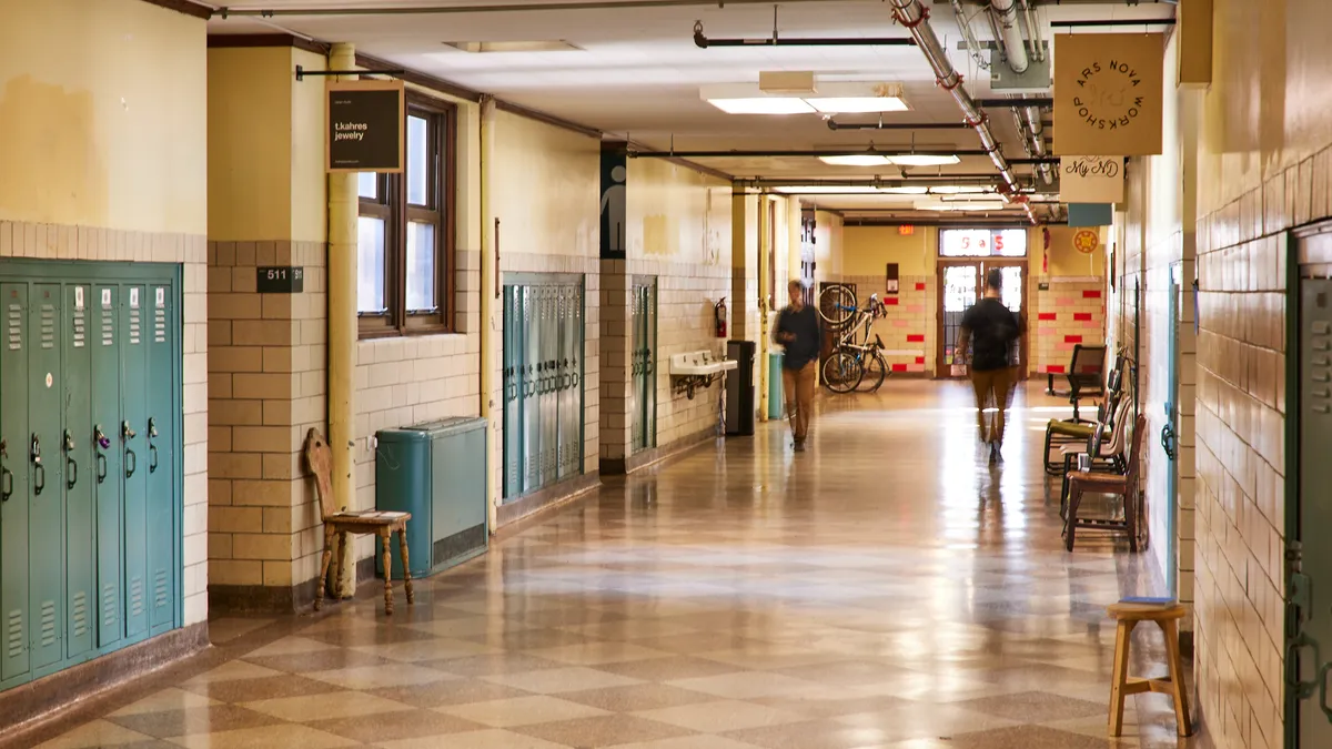 Two people walk in a hallway with lockers on the walls and signage by each door in the hallway.