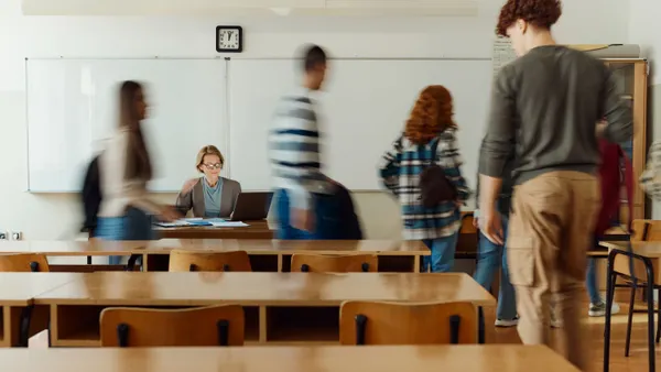 A teacher sits a desk while students stand up and leave the classroom.