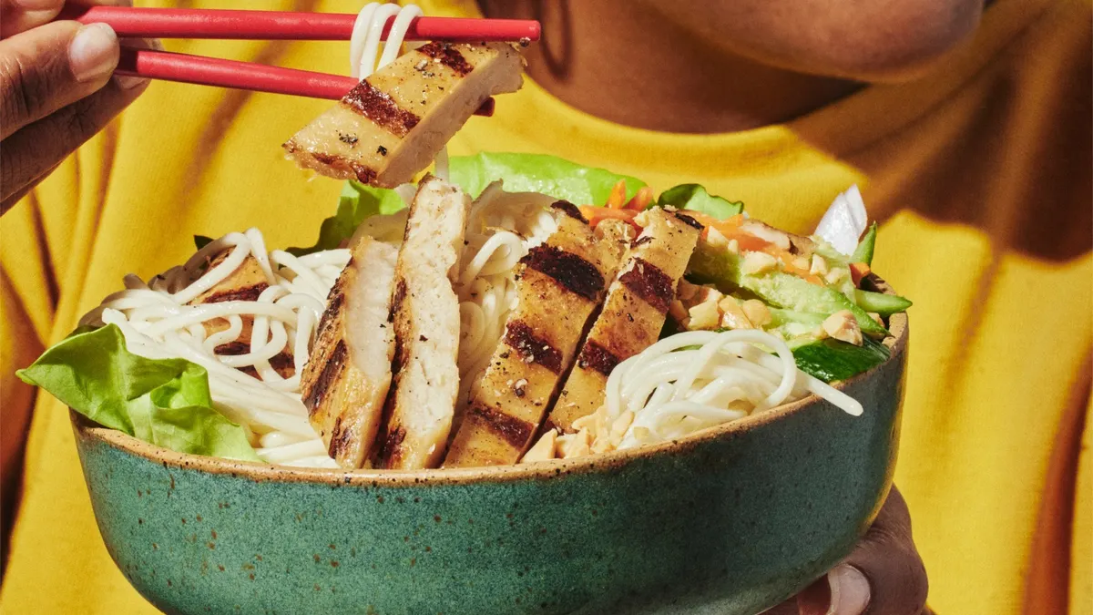 A person in a yellow shirt uses red chopsticks to pick up grilled cultivated chicken and ramen noodles in a green bowl.