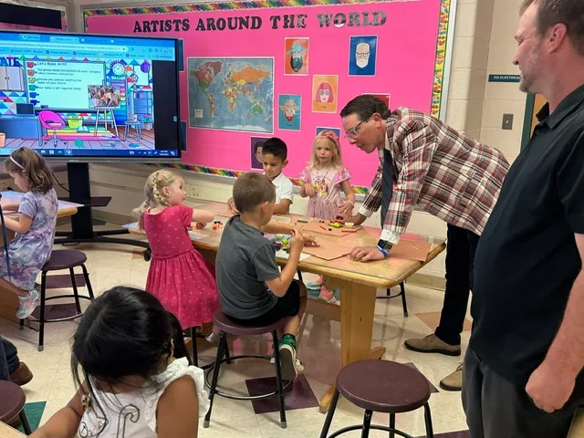 A man superintendent in a plaid jacket stands at a table in an art class to greet students on the first day of school.