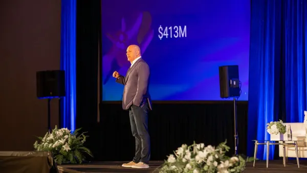 A man stands on a stage with a blue screen with the number $413 million behind it.