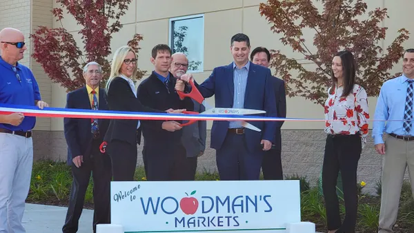 People stand outside a building during a ribbon-cutting ceremony. Several individuals hold oversized ceremonial scissors cutting a red, white, and blue ribbon. A sign in the foreground reads, “Welcome to Woodman’s Markets Lakemoor.”