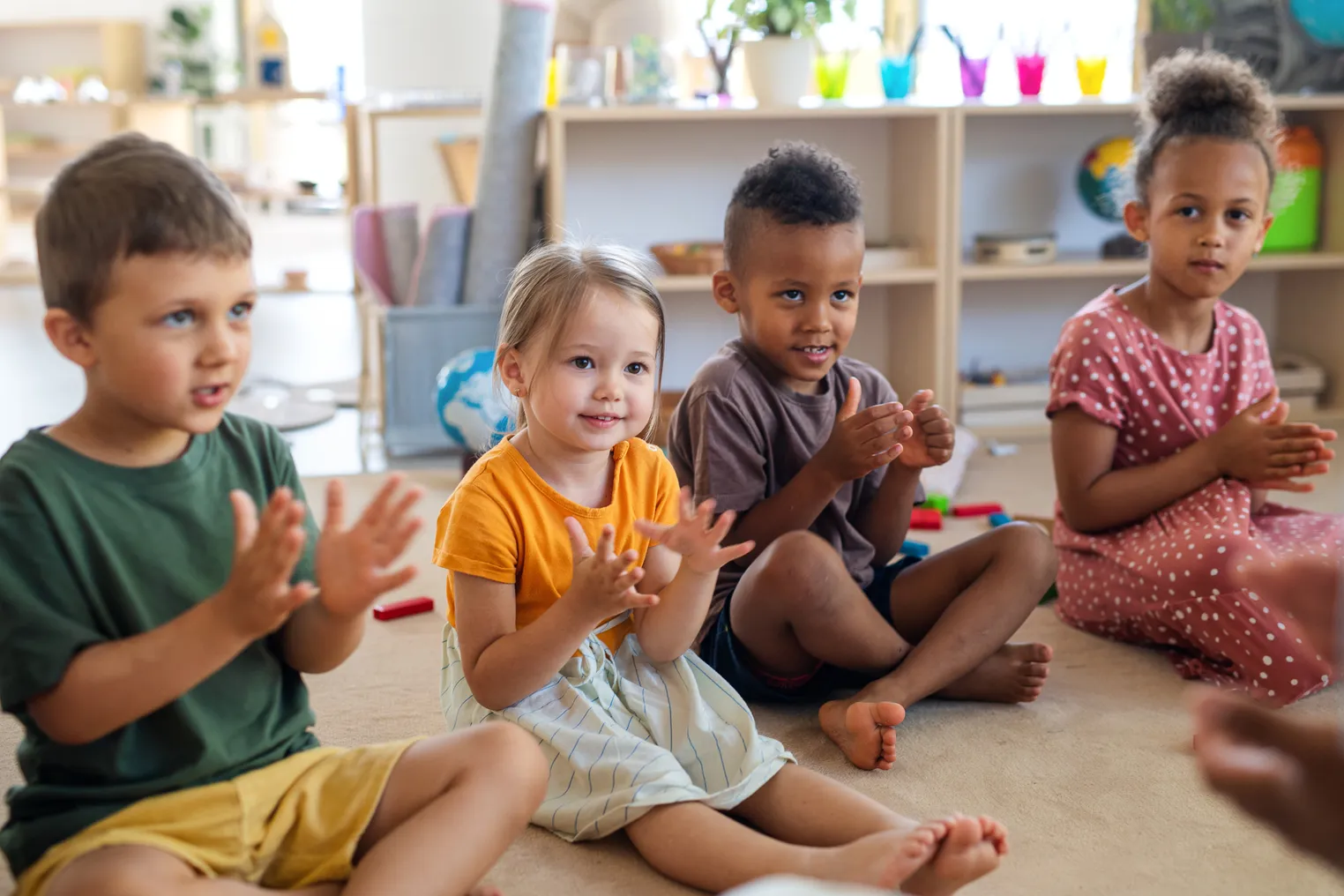 Four young children are sitting on the floor in a classroom. They are clapping