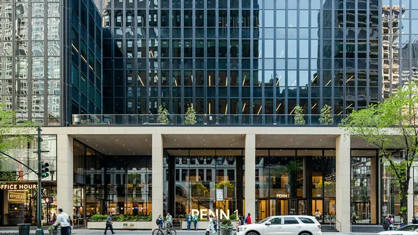 A large glass-covered building looms in the foreground on a busy street as people walk in and out of frame below and in front of it.