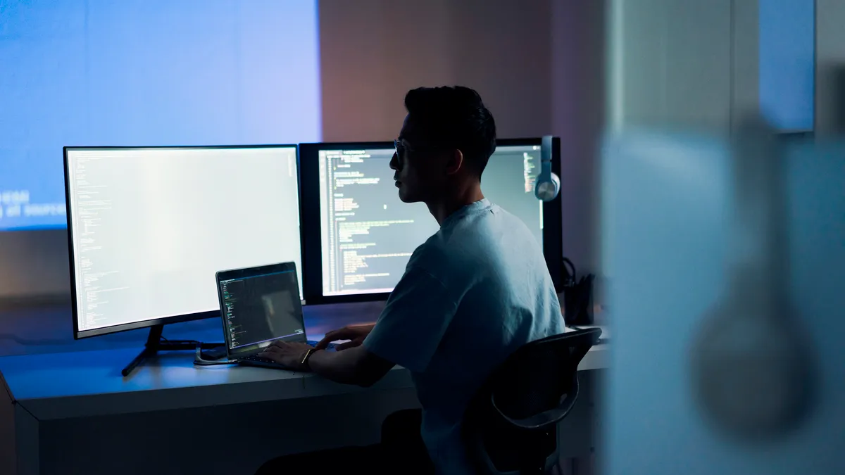 A man sits in front of a computer with lines of code upon it. The room is dark.