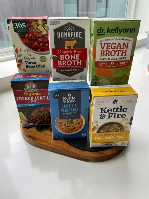 Six rectangular cartons stacked three across containing broths and soups. Top row: organic three bean chili; organic beef bone broth; vegan broth. Bottom row: organic french lentil soup; lentil &amp; vegetable hearty soup; chicken noodle bone broth soup.