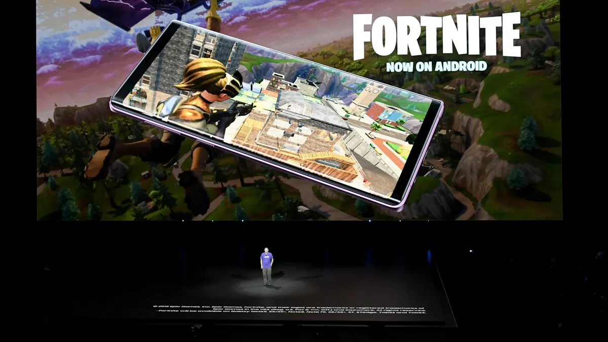 A large screen depicting the game Fortnite on a mobile phone. Standing below the screen in a a man in a purple shirt.