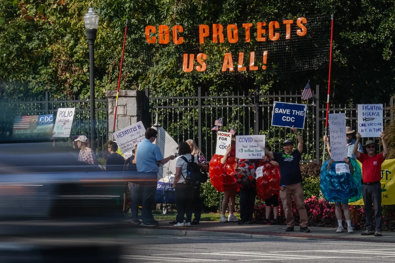 'CDC saves us all' is displayed in orange letters above an iron fence in front of green shrubbery with a stone post to the left and a lamppost to the left of the stone post. People stand in front of the fence with multiple signs, some held up, including a blue one with white letters reading ‘save the CDC.’