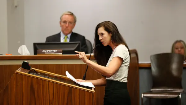 A person is standing at a podium speaking into a microphone. Another person can be seen sitting to the side of the speaker listening.