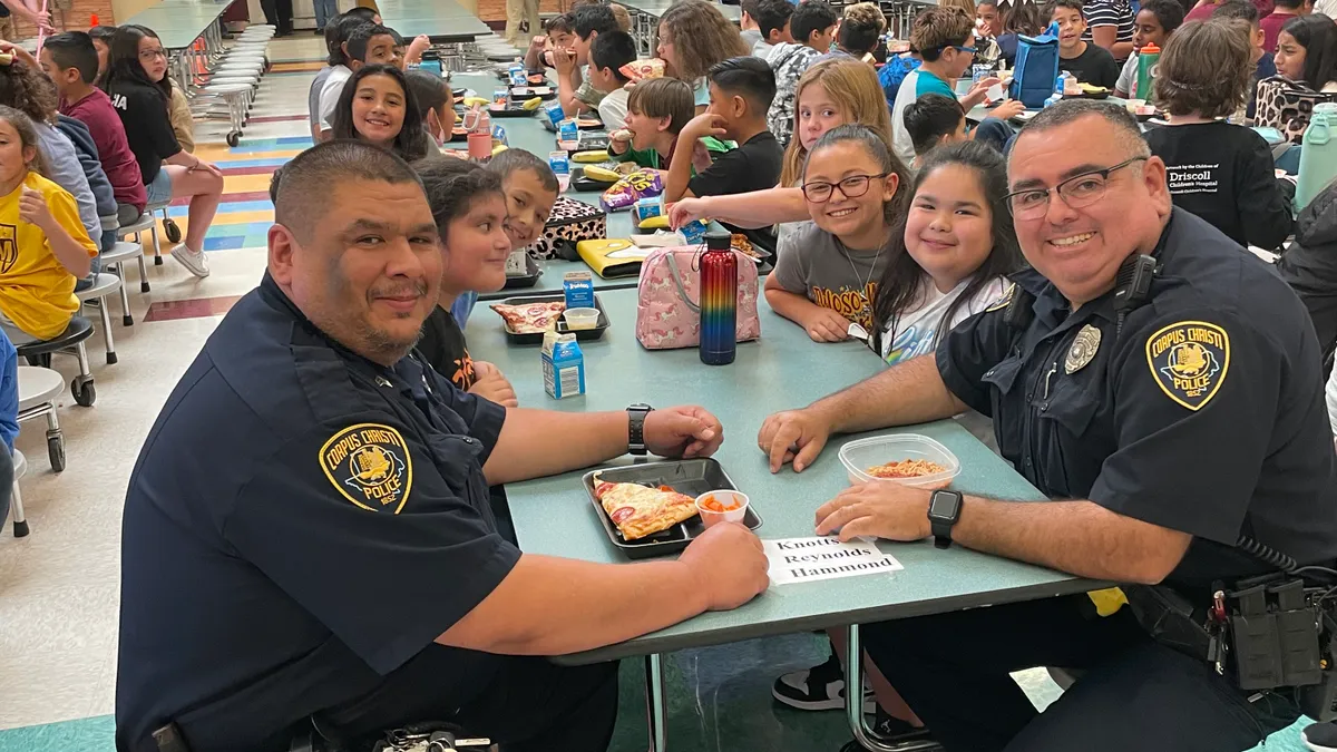 Two uniformed police officers sit in a school cafeteria at a table with students.