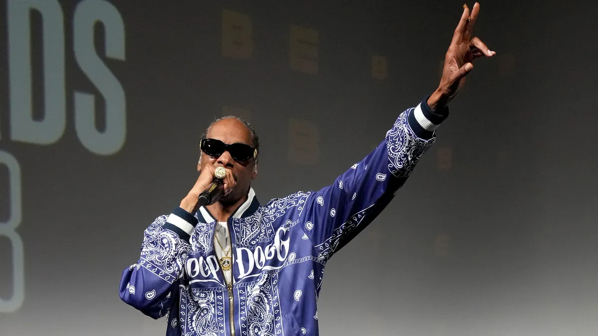 Snoop Dogg speaks into a microphone and raises his hand.