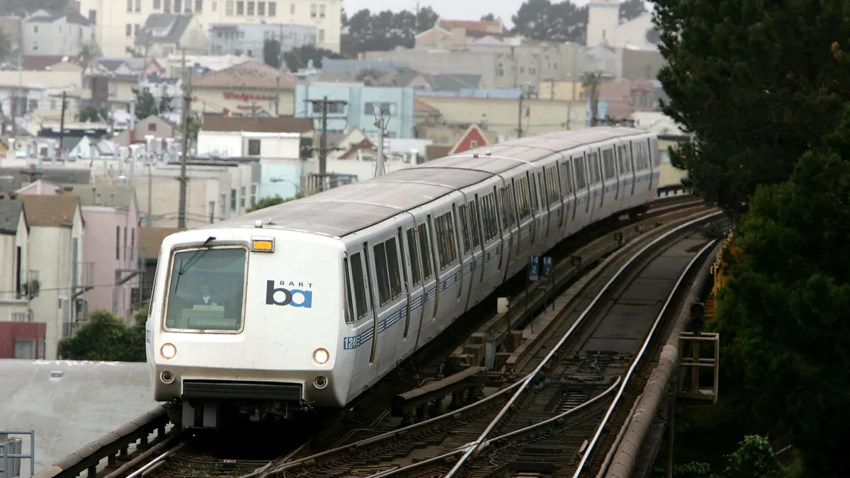 A head-on view of a Bay Area Rapid Transit, train pases residential and commercial building on an elevated track.