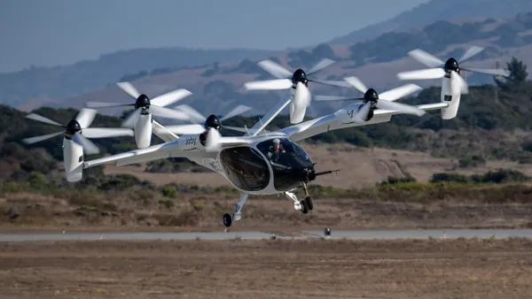 An aircraft with multiple propellers aloft over a desert with mountains in the background.