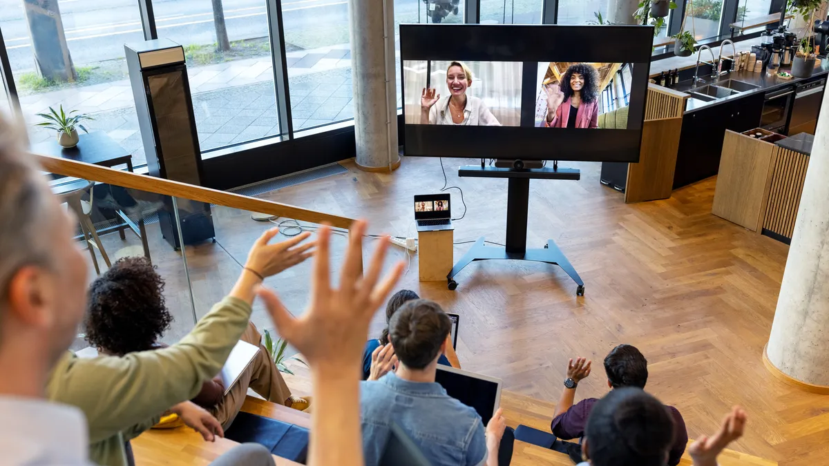 A group of employees waves to two co-workers on a video monitor.