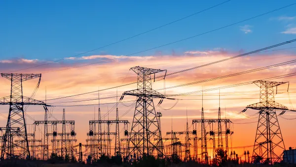 High-voltage power lines and transmission towers silhouetted against a colorful sunset sky, representing energy infrastructure and the transition towards renewable energy sources.