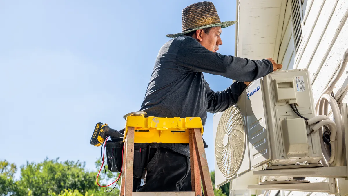 A person on a ladder reaches over to repair an air conditioning unit on a house