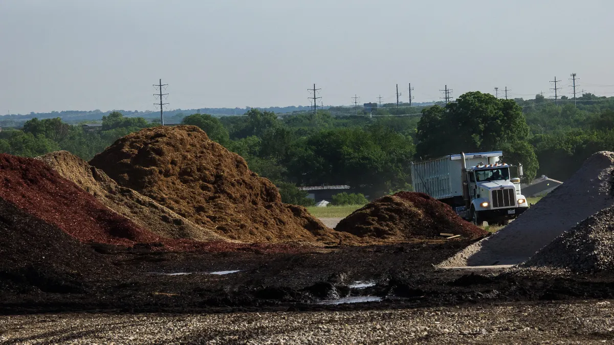 Outdoor industrial scene showing large mounds of brown, red, and dark mulch or organic material in a dirt and gravel lot. A white dump truck is partially visible behind the piles, with trees and telephone poles in the background under a cloudy sky. The ground in the foreground is muddy with scattered puddles.