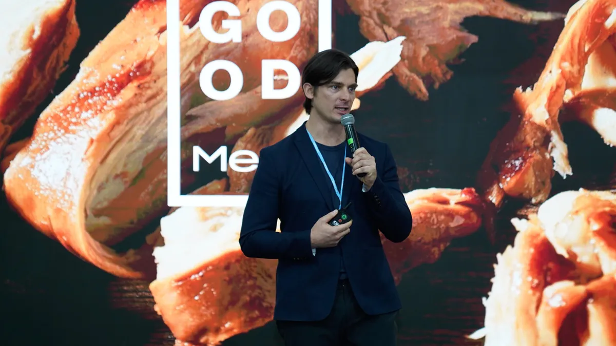 Josh Tetrick speaks into a microphone standing in front of a wall-sized image featuring pieces of Good Meat chicken.