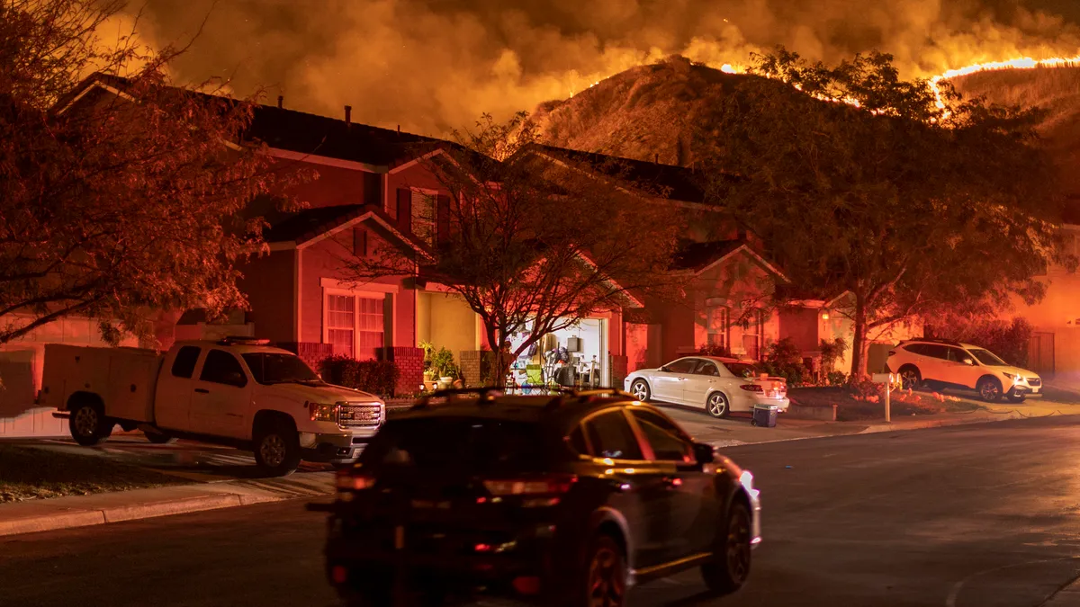 Houses and parked cars along a street lit with an orange glow. An open, brightly-lit garage at center. Trees, smoke and a line of wildfire on hills in the background.