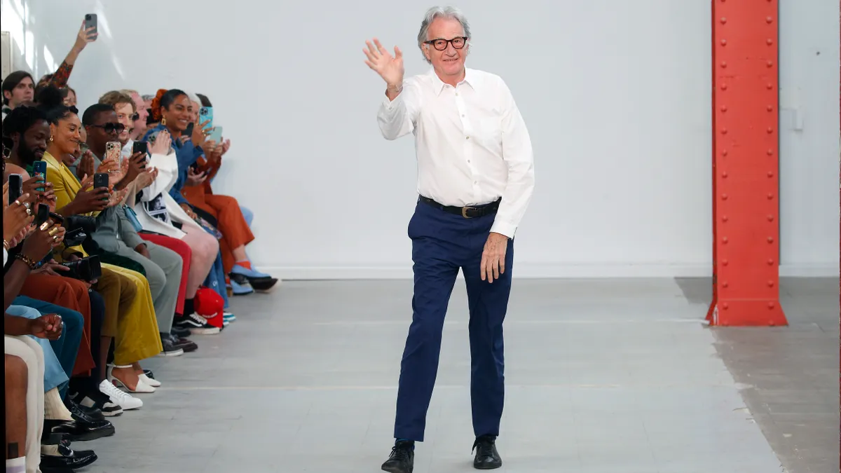 A person in a white shirt and dark pants stands on a white runway and waves at a crowd applauding from the sidelines.