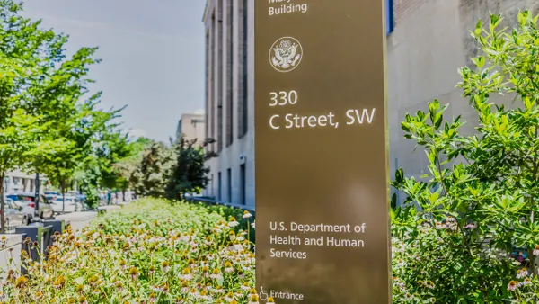 A metal freestanding sign that reads, "Mary E Switzer Building, 330 C Street, SW, U.S. Department of Health and Human Services, Entrance" is surrounded by a brush with a building in the background. 