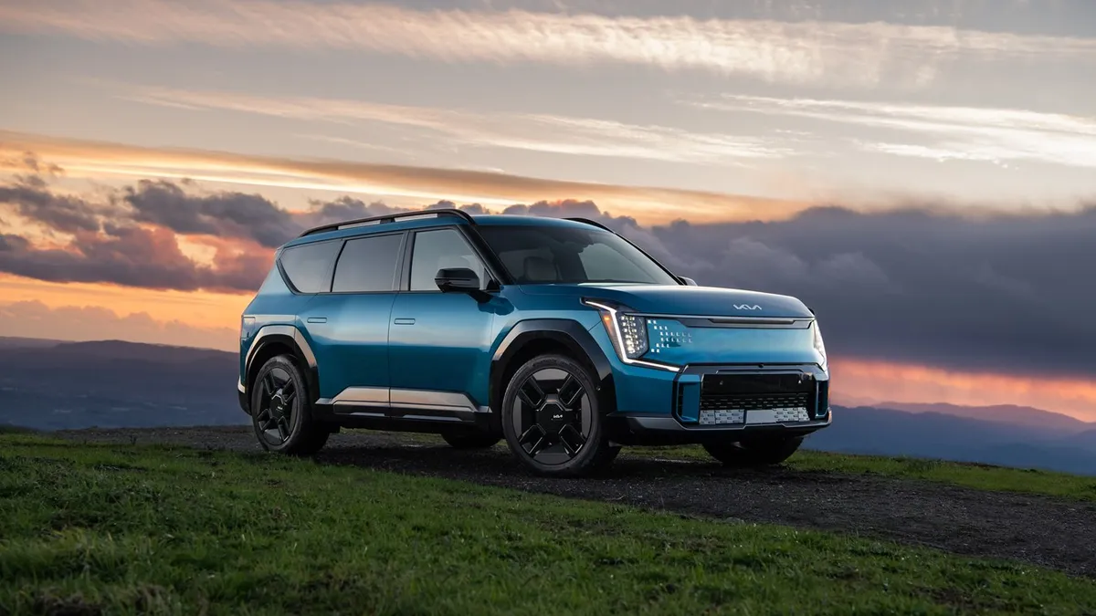 A blue Kia EV9 SUV parked outdoors with a setting sun and hills in the background.