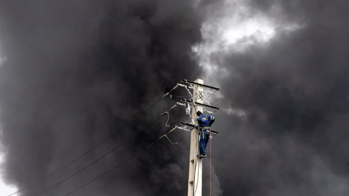 An engineer repairs an electricity pole as smoke billows after overnight airstrikes on oil depots on March 8, 2026 in Tehran, Iran.