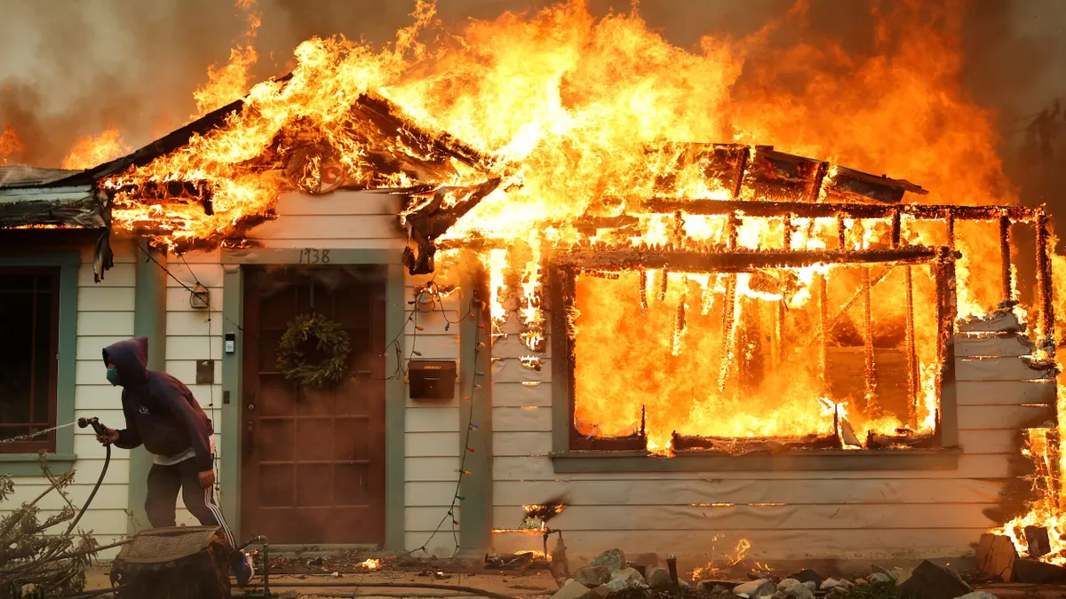 A person with a garden hose in front of a house in flames.