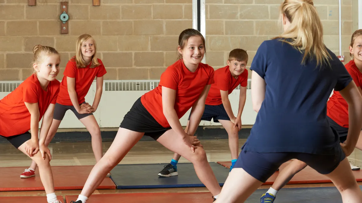 Students in physical education class face an adult while stretching in a gym. Students are wearing same red shirt and dark shorts.