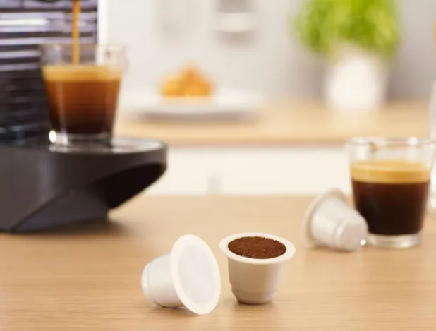 Two fiber-based coffee pods sit on a table in the foreground while an appliance brews coffee in the background.