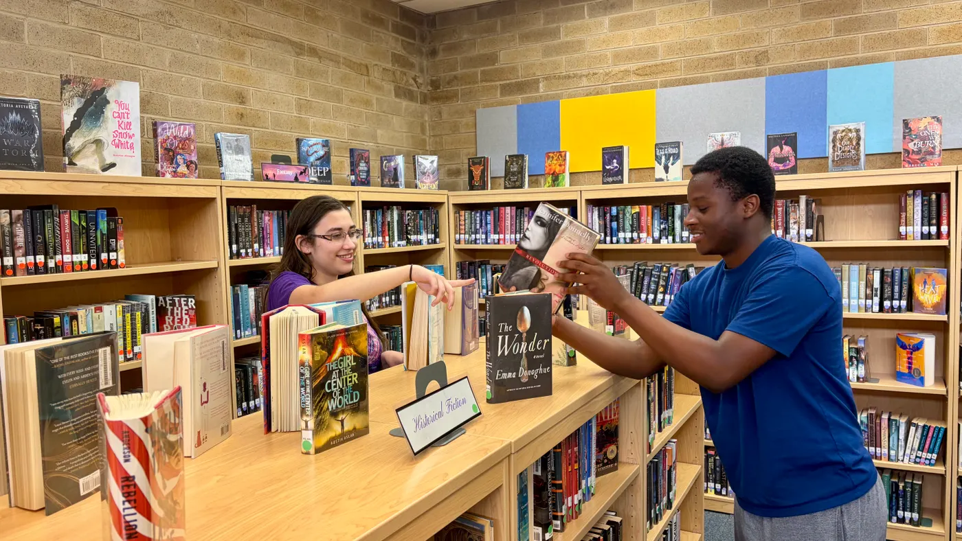 Two students are in a school library near bookselves and are passing a book over the bookshelf