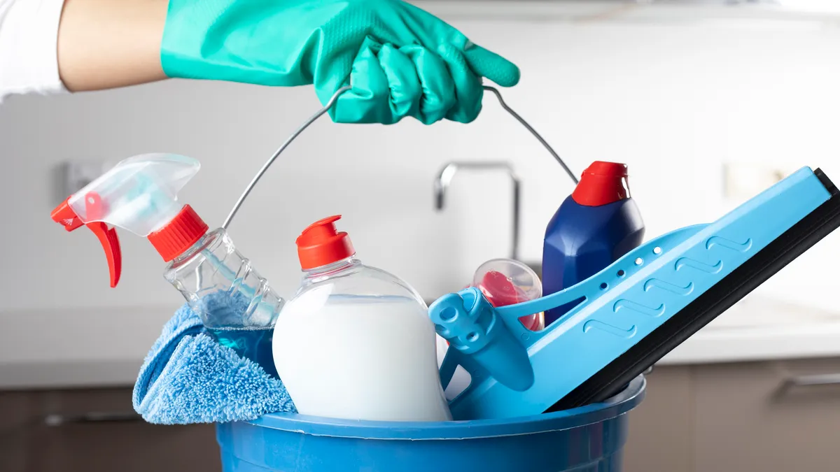 A teal color gloved hand holding a blue bucket of cleaning supplies with a white-grayish background.