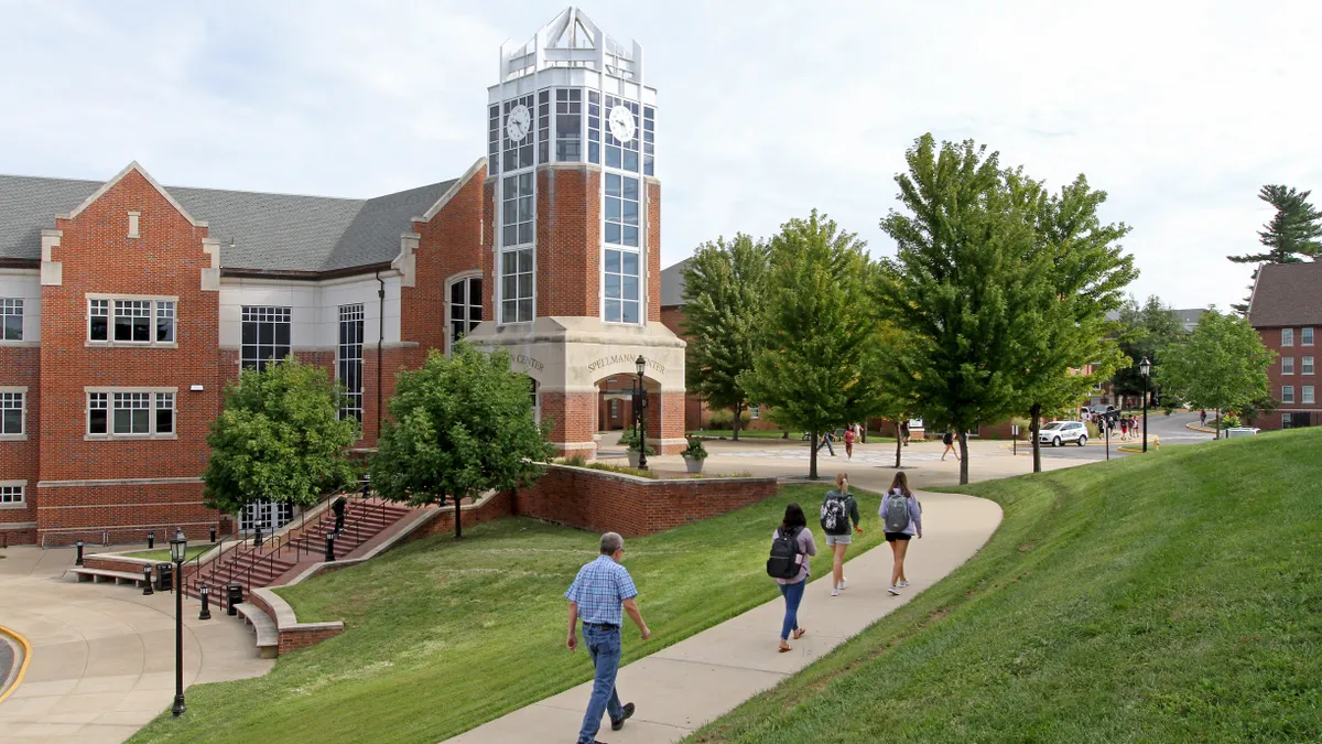 Four people walk on a sidewalk in the foreground with a sprawling green college campus in the background.